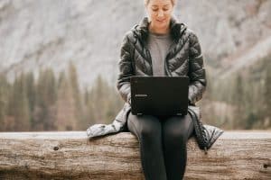Photo of woman working on laptop sitting outside on a large log on a hillside